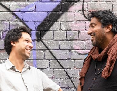 Two men smiling and laughing against the backdrop of a brick wall covered in urban art.