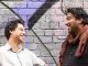 Two men smiling and laughing against the backdrop of a brick wall covered in urban art.